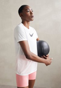 Camisa atlética de manga corta blanca con logo gris oscuro, combinada con pantalones cortos rosas, sosteniendo una bola de peso negra contra un fondo neutral.
