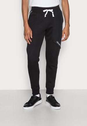 Mann trägt schwarze Jogginghosen mit weißem Kordelzug, seitlicher Klappentasche mit Logo, weißen Socken und schwarzen Turnschuhen auf grauem Boden.