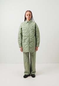 Jachetă matlasată verde deschis, cu închidere cu nasturi și două buzunare pe piept, asortată cu pantaloni matlasați de aceeași nuanță și pantofi negri.