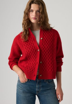 Young woman with wavy hair wearing a red textured buttoned cardigan over a black and white striped top and blue jeans, standing with one hand in pocket.