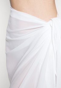 Envoltura de tela blanca transparente con un detalle de nudo retorcido en un lado, con una textura suave y un diseño drapeado que revela parcialmente la zona media.