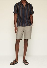 Camisa de manga corta con rayas verticales en azul marino y marrón, combinada con pantalones cortos gris claro y sandalias negras.