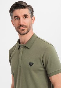 Olivgrön polo skjorta i bomullsmix, med en svart triangel-logotyp på vänster bröst, klassisk krage och korta ärmar.