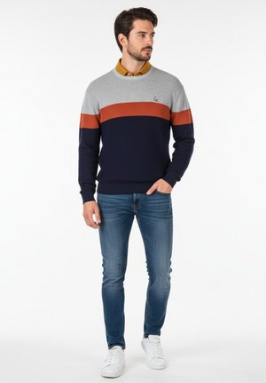 Suéter bloqueado en color azul marino, gris y naranja con textura acanalada, combinado con jeans ajustados azul y zapatillas blancas.