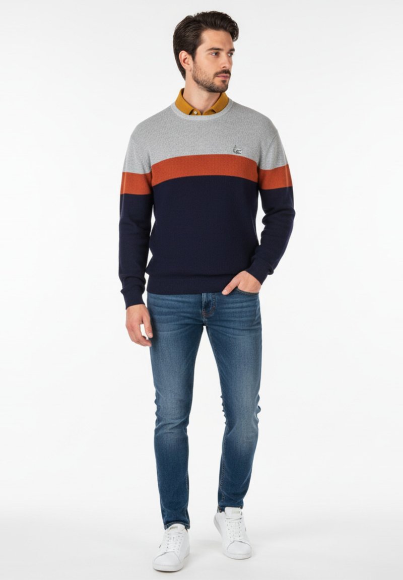 Suéter bloqueado en color azul marino, gris y naranja con textura acanalada, combinado con jeans ajustados azul y zapatillas blancas.