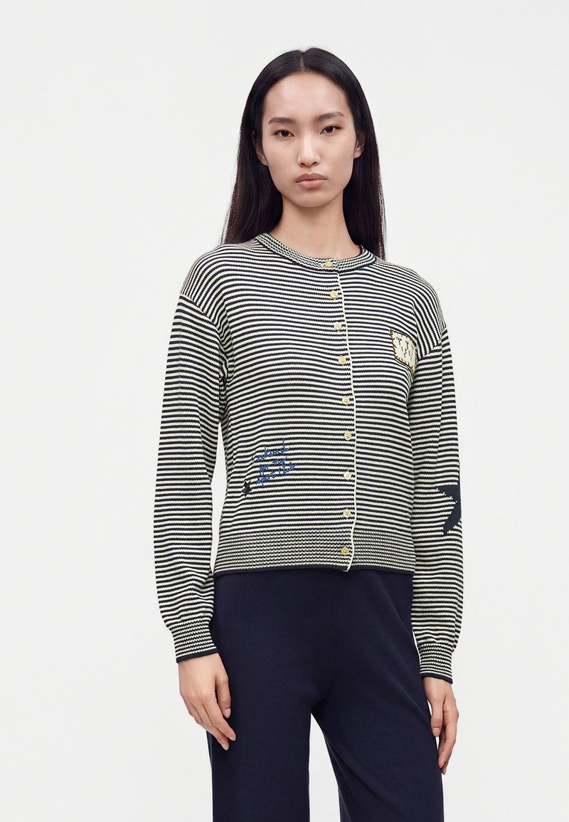 Femme aux longs cheveux noirs portant un cardigan rayé noir et blanc boutonné avec de petites pièces et un pantalon foncé sur fond blanc.