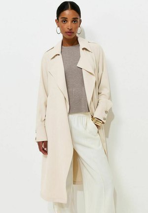 Donna che indossa un trench beige, un maglione taupe, pantaloni bianchi, braccialetti dorati e orecchini a cerchio, in piedi davanti a uno sfondo bianco.