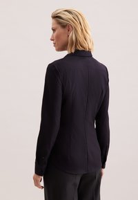 Kobieta z krótkimi blond włosami zwrócona w lewo, ubrana w dopasowaną czarną bluzkę z długim rękawem i czarne spodnie na jednolitym tle.