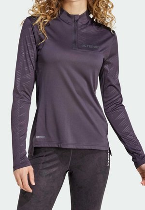 Kvinde iført mørkelilla langærmet atletiktop med kvart lynlås og matchende leggings, begge med "TERREX" og "AEROREADY" logoer.