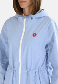 Veste bleu clair en nylon avec une fermeture éclair blanche, une ceinture élastique et un logo circulaire rouge sur la poitrine. Comprend une capuche.