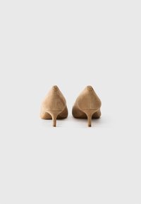 Pair of beige suede kitten heels seen from the back on a plain white background, showing small round gold logos on each heel.