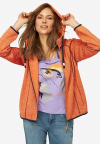 Giacca arancione con texture, zip e cappuccio, caratterizzata da dettagli neri e due tasche laterali. Indossata sopra una maglietta viola con una grafica di un animale.