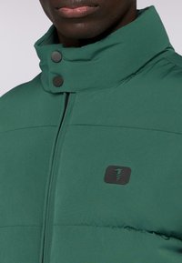 Zelená vyp paddingová bunda s vysokým límcem, černými druky a malým černým logem na hrudi, s hladkou texturou a strukturovaným designem.