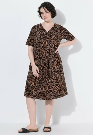 Kvinde med kort krøllet hår står op ad en hvid væg iført en brun leopardmønstret kjole og sorte slippers, hånd i siden.