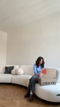 Cardboard box with Zalando logo, placed on a curved white sofa. The setting features a black and white patterned cushion and light wood flooring.