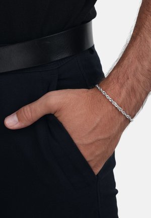 Pulsera de cadena de plata con un diseño de eslabones intrincado, llevada en una muñeca parcialmente visible dentro de un bolsillo, combinada con un cinturón negro y ropa.