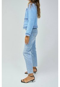 Ljusblå texturerad jacka med fransar, i kombination med ljusblå raka jeans och svarta sandaler med remmar.