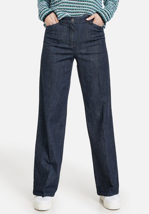 Persona che indossa jeans a gamba larga blu scuro con le mani nelle tasche e sneakers bianche con zeppa, in piedi contro uno sfondo uniforme.