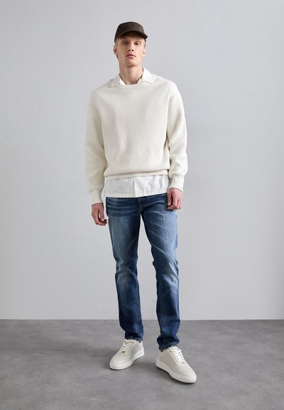 Pulover ribbed de culoare crem, purtat peste o cămașă albă, asortat cu blugi albaștri decolorati și adidași albi cu accente texturate.