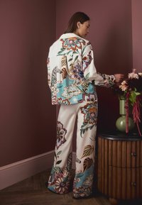 Woman in floral-patterned white shirt and pants arranges flowers in green vase on wooden cabinet against maroon wall.