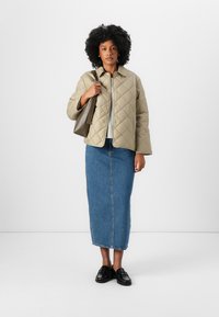 Veste matelassée beige avec un devant à boutons, pull gris en dessous, associée à une longue jupe en denim bleu et des mocassins noirs. Tient un sac marron.