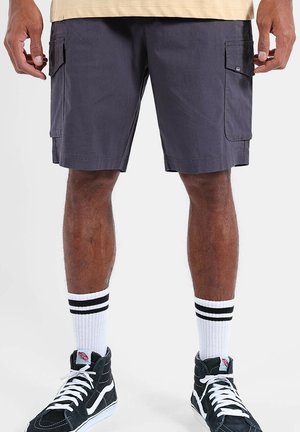 Homme portant un short cargo gris foncé, des chaussettes blanches à rayures noires et des baskets montantes noires avec des lacets blancs, debout.