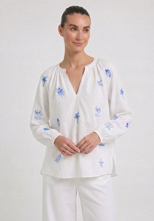 Frau trägt eine weiße Bluse mit langen Ärmeln und blauen, bestickten maritimen Motiven, kombiniert mit weißen Hosen, steht vor einem einfarbigen Hintergrund.