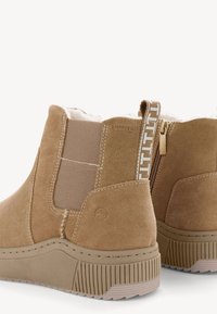 Bottines en suede marron avec des panneaux élastiques sur les côtés, semelle en caoutchouc texturée et une tirette décorative avec un logo. Doublure douce pour le confort.