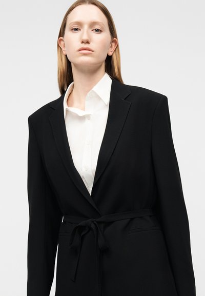 Γυναίκα με μακριά ίσια μαλλιά, που φοράει λευκό πουκάμισο με γιακά και υπερμεγέθη μαύρο σακάκι δεμένο στη μέση, σε ουδέτερο φόντο.