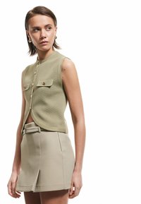 Ljusgrön, ärmlös topp med en texturerad yta, knappar framtill och två bröstfickor; matchad med en beige midjeförsedd kjol.