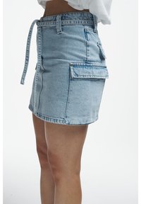 Denim mini skirt with cargo pockets and a matching denim belt worn with a white top, shown from the side on a person.