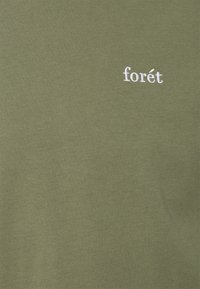 Oliivinvihreä puuvillainen t-paita, jossa vasemmalla valkoisella kirjailtu teksti "forét". Sileä pinta, yksinkertainen muotoilu ja pyöreä pääntie.