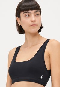 Sutien sport din bumbac negru, cu decolteu în formă de cerc și benzi ribbate de susținere. Prezentă un logo alb pe partea stângă. Textură netedă și elastică.