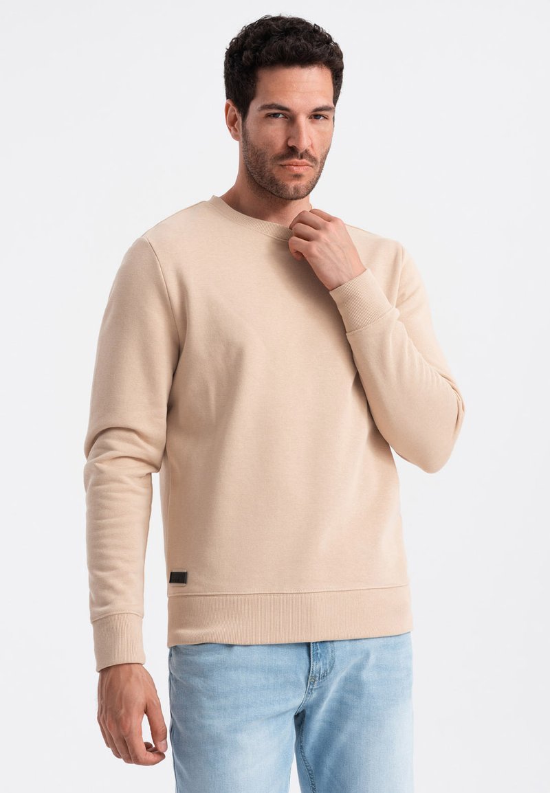 Sudadera beige de cuello redondo con mangas largas, puños y dobladillo acanalados. Presenta una pequeña etiqueta de la marca en negro en la parte inferior izquierda.
