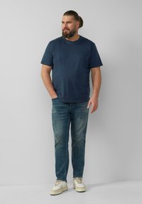 T-shirt in cotone blu navy, vestibilità comoda, abbinato a jeans blu sbiaditi e sneaker crema con dettagli chiari. In piedi contro uno sfondo neutro.