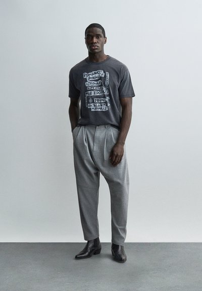 Άνδρας στέκεται μπροστά σε απλό τοίχο φορώντας σκούρο μπλουζάκι με στάμπα και κείμενο, ανοιχτό γκρι παντελόνι με πιέτες και μαύρες μπότες.