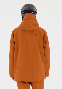 Orangene wasserdichte Jacke mit großer Kapuze, Reißverschluss-Rückenfach und geräumigen Ärmeln. Strukturierter Stoff geeignet für Outdoor-Aktivitäten.