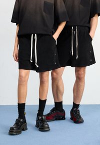 Zwarte textuurshorts met witte trekkoord, voorzien van zijzakken en metalen accenten, gecombineerd met oversized zwarte gradient T-shirts en contrasterend schoeisel.