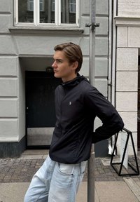 Noor mees musta jope ja helesiniste teksadega toetumas linna kõnniteel asuvale metallpostile halli hoone lähedal.