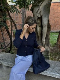 Marinblå kabelstickad kofta med runda knappar, vit flödande kjol och en mörk denim axelremsväska på en stenbänk omgiven av grönska och tegelvägg.