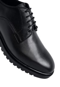Zapato de vestir de cuero negro con puntera redonda, cierre con cordones y suela de goma texturizada sobre un fondo blanco.
