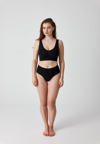 Vrouw staat blootsvoets, draagt een zwarte sportbeha en hoog uitgesneden ondergoed tegen een effen lichte achtergrond.