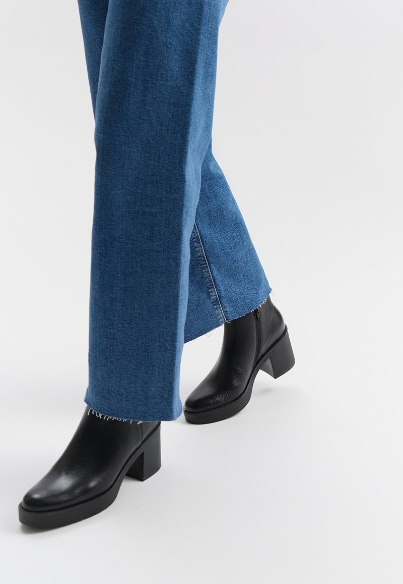 Personne portant un jean en denim bleu et des bottines en cuir noir à talons carrés sur un fond de sol blanc.
