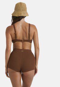 Brauner gerippter Bikini-Oberteil mit verstellbaren Trägern und goldenen Beschlägen, kombiniert mit hochgeschnittenen braunen gerippten Shorts. Geflochtene Strohhut im Bucket-Stil.