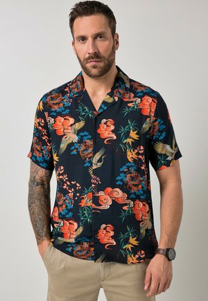 Mann mit Bart, der ein kurzärmeliges schwarzes Hemd mit orangefarbenen, blauen und grünen Blumen- und Vogelmustern, beige Hose und eine Armbanduhr trägt.