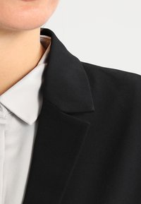 Černý blazer s výstřihem do V, hladkou strukturou, nošený přes světle šedou košili. Detailní záběr ukazuje detaily límce a povrchovou úpravu látky.