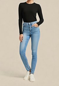Femme portant un haut à manches longues noir, un jean skinny taille haute bleu clair, des baskets blanches, avec une main dans la poche, sur un fond beige.