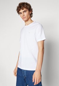 White cotton t-shirt with a round neck, short sleeves, and a small, stitched logo on the left chest. Paired with blue denim jeans.