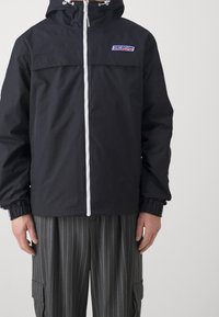 Czarna wodoodporna kurtka z kapturem, białym zamkiem błyskawicznym i wyszywanym logo na piersi, wyposażona w elastyczne mankiety i luźny krój.