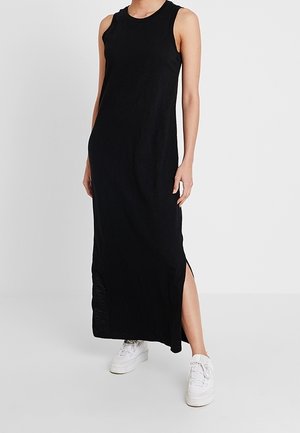 Kvinde iført en sort maxi-kjole uden ærmer med sideslids og hvide sneakers, stående mod en ensfarvet lys baggrund.